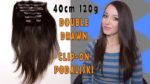 Žena s dugom smeđom kosom prikazuje ekstenzije za kosu Double Drawn Clip-On, duljine 40 cm i težine 120 g.